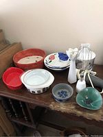 Photo showing multiple kitchen items including a red pottery bowl, floral decorated dish, Pyrex casserole, red ceramic dish, two small Italian pottery bowls, and immersion blender parts.