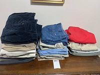 Stack of assorted mens pants and shorts including corduroy, denim, and khaki styles in various colors