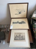 Three framed prints stacked on a table and leaning, showing titles and scenes with people, buildings, and natural settings.