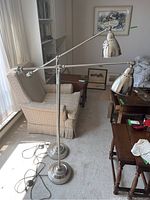 Two metal adjustable arm floor lamps standing side by side in a living room. Both have weighted round bases and chrome finish, with dome-shaped shades featuring ventilation holes.