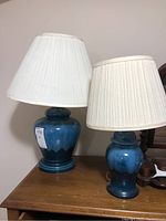 Pair of two blue ceramic lamps with white fabric lampshades shown together on wooden surface. Lamps differ in height around 25 and 20 inches. Both lamps exhibit loose top portions as noted.