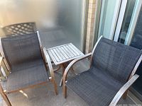 Two wooden frame patio chairs with black canvas seating alongside a small wooden end table with a slatted top on a balcony or patio floor.