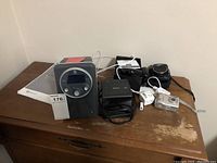 Photo showing lot with Boston Horizon Solo radio manual, Sony alarm clock, Canon PowerShot SX150IS camera, Nikon Coolpix 4600 camera and Widex devices on wooden table.