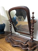 Front view of solid wood shaving mirror with curved base, ornate twisted wooden supports, and rectangular mirror with rounded top corners placed on a wooden surface next to lamps.