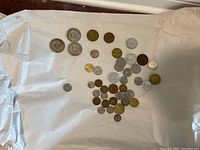 Wide shot showing the assortment of approximately 40 coins including the three large silver Venezuelan coins and other smaller mixed metal coins arranged on white paper.