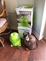 Two Crane ultrasonic cool mist humidifiers, frog and owl shaped, with boxes displayed beside a chair on wooden floor.