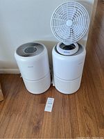 Two white Levoit air purifiers side by side with a small white Holmes table fan placed on top of one air purifier.