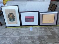 Three framed artworks placed side by side on the floor; includes an Egyptian papyrus art, a small landscape painting, and a smaller papyrus or parchment piece.