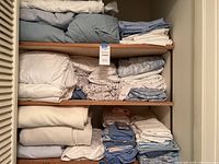 Shelf with multiple stacks of folded bed linens including white, light blue, and patterned sheets, pillowcases, and comforters.
