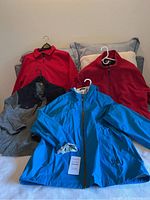 Four men's jackets displayed on a bed: bright blue windbreaker, two red fleece jackets, and one gray/black fleece jacket.