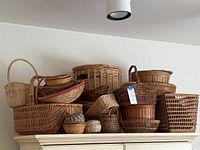 Photo showing assortment of woven baskets arranged on top of a cabinet. Various sizes and styles visible including round, oval, rectangular, handled, and shallow weaving styles