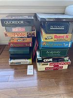Two stacks of assorted used board games placed on the floor including Monopoly, Clue, Scrabble, Settlers, and others.