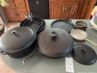 Full view of cast iron and non-cast cookware grouped on table - large cast iron skillet with lid, kettle, griddle, smaller skillets and pots