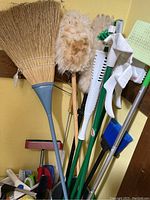 Photo showing multiple cleaning tools including brooms, dust mops, brushes, and mop handles arranged in a corner.