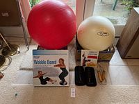 Photo showing red and off-white stability balls, Wobble Board in box, Complete Stretching booklet, ankle weights, and jump rope arranged on floor.