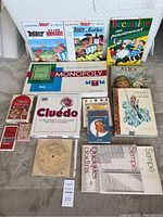 Full lot items on carpet including two Asterix comic books, Bécassine au pensionnat book, Monopoly game, Cluedo game, Alice book, Grimm Contes book, and various other books and a CD.