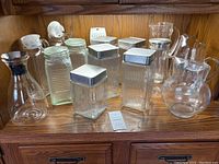 Wide angle shot of the entire collection of glass storage containers, pitchers, and jars on wooden shelf, showing their relative sizes and shapes. Some containers have metallic or plastic lids; all are clear glass.