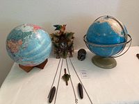 Two vintage globes, a carved wooden cuckoo clock with weights and pendulum, and a small clay medieval helmet placed on a table against a wall.