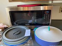 Samsung stainless steel microwave with digital display showing time 1:45, grey collapsible steamer basket, blue bowl with white lid and green handle, and red silicone mat.