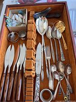 Photo of wooden handled knives, large serving spoons, carved scoop, and some smaller cutlery pieces in a wooden tray with velvet slots