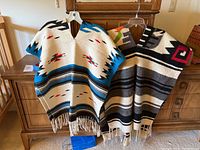 Two ponchos displayed on hangers showing different Native American pattern designs and bottom fringes.