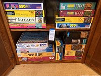 Stack of assorted boxed jigsaw puzzles on shelf includes National Geographic 1000 Sailing Ships, 1500 Super Puzzle, Cobble Hill 1000 piece, Golden Gallery 550 Peacock Pride and more