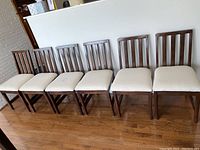 Photo showing all six wooden dining chairs lined up side by side, cream upholstered seats, dark wood frames.