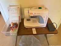 Singer Simple sewing machine on attached table with foot pedal visible. Accessories and manuals arranged in a clear plastic bin beside the machine.