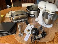 View of all appliances on a wooden table showing KitchenAid mixer, Crockpot, Cuisinart Griddler with plates, Cuisinart hand blender parts and manual