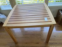 Full view of a square teak table with slatted top and sturdy legs set on a wooden floor.