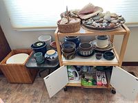 Wooden rolling rack loaded with assorted pots and baskets with shells. Open cabinet doors show some garden supplies and tools below.