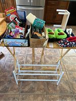 Two wooden TV trays filled with assorted household items and slippers positioned behind a white metal shoe rack on tiled floor.