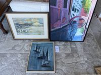 Three framed vintage paintings arranged on a carpeted floor. One has a cloudy sky with sailboats, another a textured sailboat scene, and the third a Venice canal.