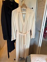 Front view of cream wool gabardine wrap coat with belt and dark colored coats behind it.