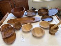 Photo showing the full collection of wooden serving pieces arranged on a white cloth backdrop including bowls, plates, tray, and mortar and pestle.
