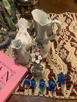 Group of ceramic vases and pitchers with floral motifs, small blue glass animal figurines arranged on a crocheted cloth along with a pink book.