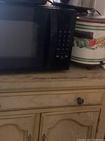 Black microwave oven with numeric keypad on the right side, placed on a worn wooden dresser next to a crock pot with a vegetable pattern.