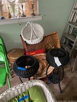Photo showing multiple woven baskets of various shapes and colors stacked and adjacent to two black wicker plant stands with metal legs.