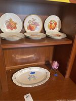 Shelf with nine china plates featuring fruit designs, a pink decorative egg on stand, and an oval platter below.