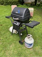 Full view of black Thermos branded single burner BBQ grill outdoors on green grass with one propane tank beside it and another under the grill. Grill has side shelf and two wheels.
