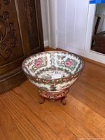 Photo of decorative bowl on ornate stand showing Asian-inspired floral and bird designs, placed on wooden floor near carved wooden furniture.