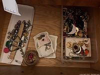 Wide view showing assorted jewelry items on wood surface: watch, bracelets, trays with pins and earrings, and trinket box.