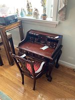 Angled view of the miniature wooden desk with many small drawers and the carved chair with red upholstered seat.
