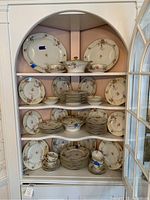 Full view of china set arranged in cabinet showing multiple shelves filled with plates, bowls, cups, and serving pieces mainly in white with floral design and gold trim.