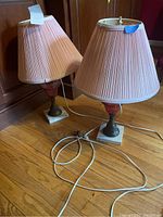 Pair of table lamps shown together on wood floor with beige pleated fabric shades and electric cords visible.