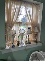 Four lamps displayed on a windowsill with curtained background including two white glass tulip style lamps and two candlestick lamps with fabric shades.