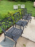 Set of four black cast aluminum patio chairs with lattice seats and decorative scrollwork backs placed on a sidewalk near greenery.