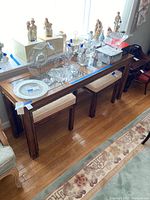 Wood sofa table with glass top paired with two matching wood benches with upholstered seats, shown in a well-lit room with floor rugs and other decorative items on top.
