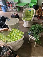 Photo showing two white wicker scoop chairs with green cushions, one wicker side table, and a wicker plant stand with faux plant in a garage setting.