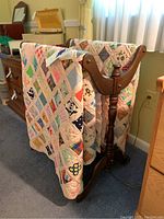 Wood quilt rack holding two patchwork style queen size quilts and a fabric valance, showing the front and side angles of the rack and fabrics.